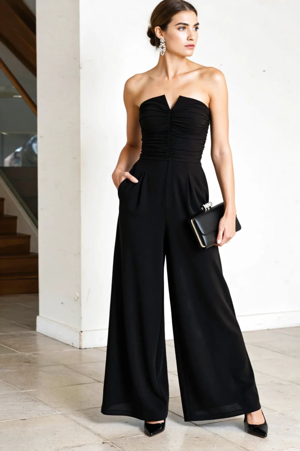Model in a strapless black crepe wide-leg jumpsuit with cinched waist, pointed-toe heels, statement earrings and a sleek clutch