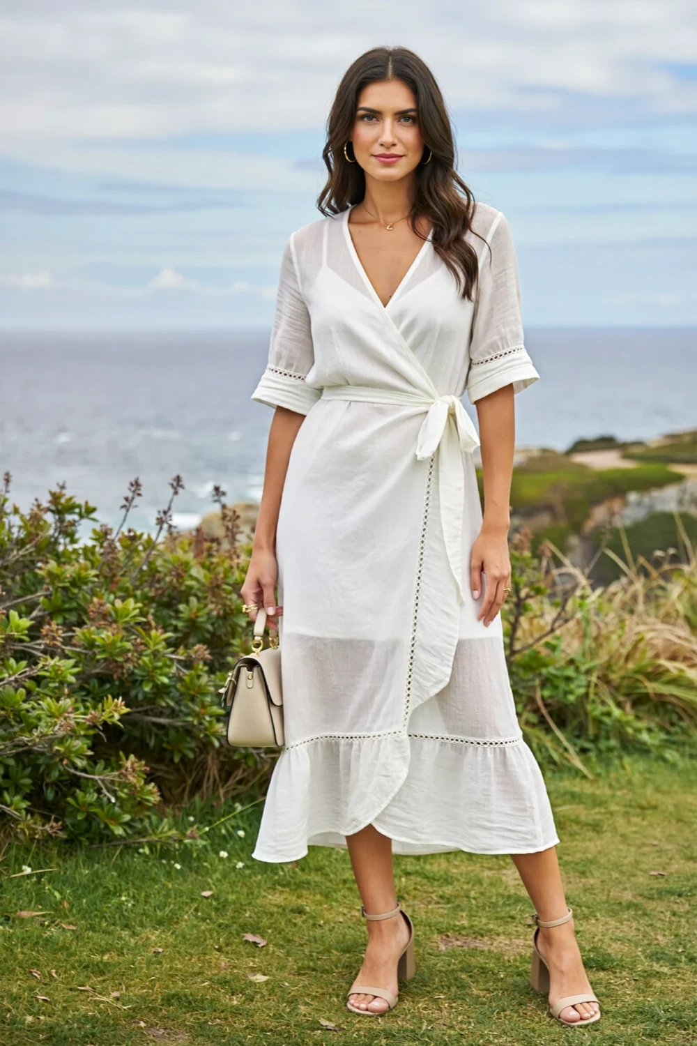 Model in a white linen wrap dress with V-neck and tie waist, styled for a beach wedding guest on grassy coastal cliffs.