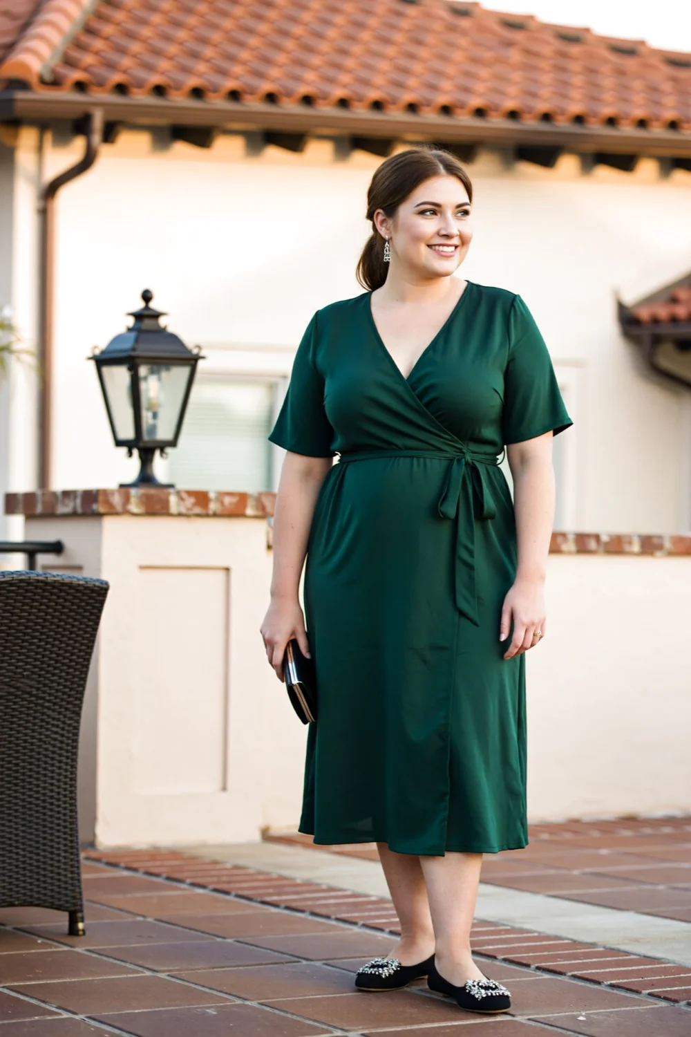 Woman wearing emerald wrap midi dress with defined waist, V-neckline and tied slim belt, clutch and jeweled flats on tile terrace