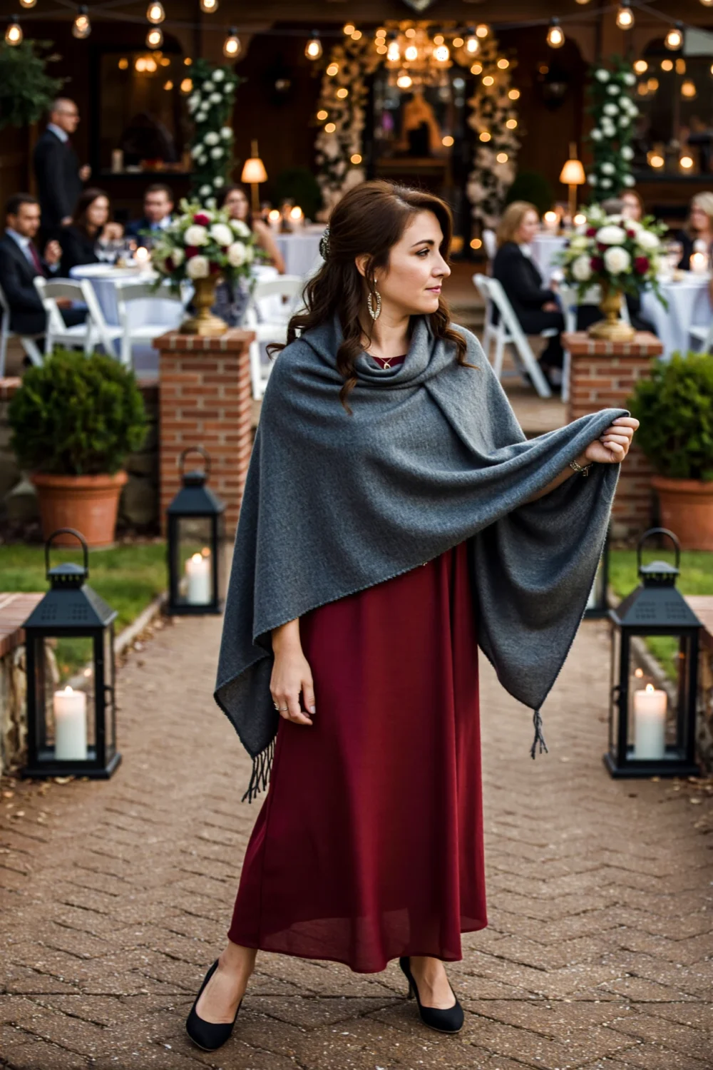 Wedding guest wearing a grey pashmina draped over a burgundy dress, an elegant coverup at a candlelit outdoor reception for warmth